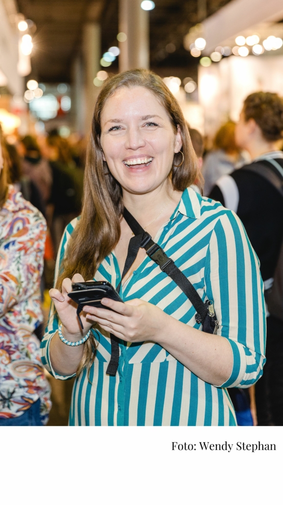 Foto von Svenja Hirsch auf der Frankfurter Buchmesse. Sie hat ein Handy in der Hand und lacht in die Kamera.